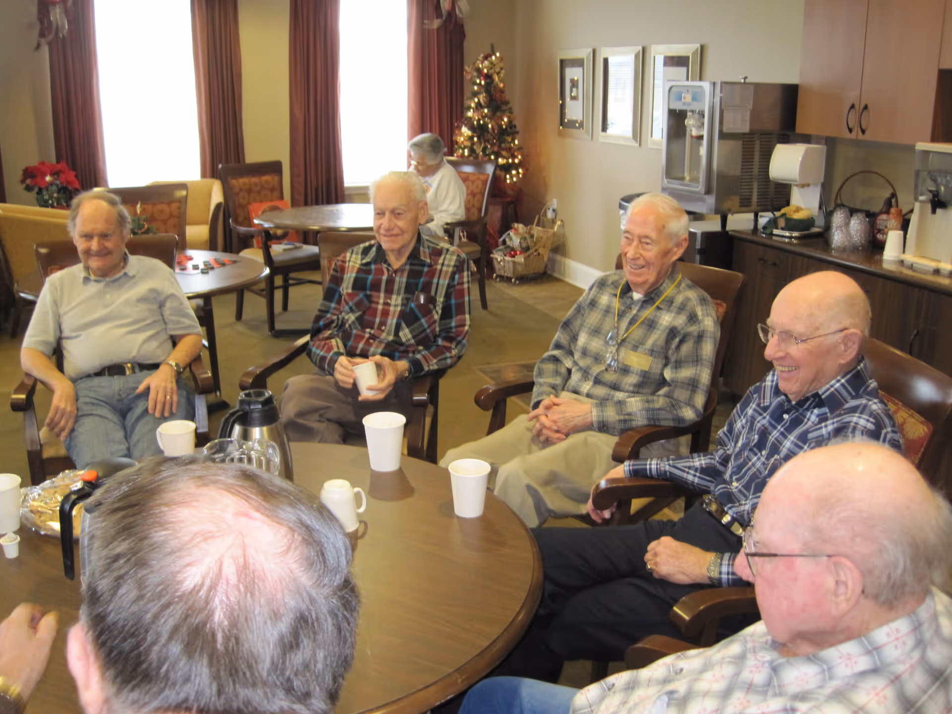 A group of elderly men sitting around a round table in a senior living community common area, smiling and enjoying each other's company. The room has large windows with curtains, a decorated Christmas tree, and a kitchenette area with a coffee machine and cups.
