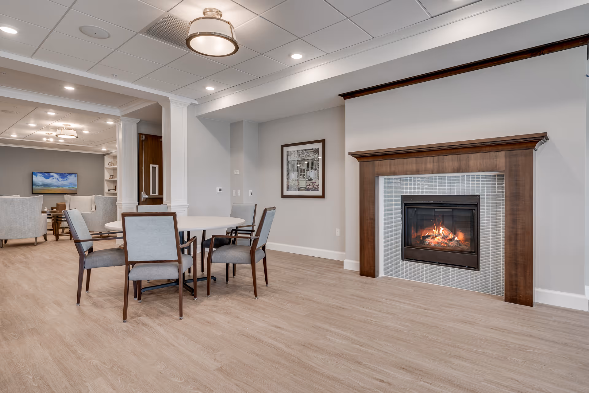 A cozy senior living common area featuring a round table with four chairs in the foreground, a lit fireplace with a wooden mantel on the right wall, and a seating area with armchairs and a TV in the background. The room has light wood flooring, neutral-colored walls, and recessed lighting in the ceiling.
