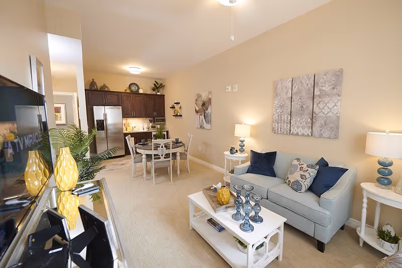A bright and cozy senior living apartment interior featuring a living room with a light blue sofa adorned with blue and patterned pillows, two side tables with blue lamps, and a white coffee table decorated with blue candle holders and a yellow vase. Adjacent to the living room is a dining area with a round table and four chairs, and a kitchen with dark wood cabinets and stainless steel appliances. The walls are painted beige, and there are decorative paintings and plants enhancing the space.