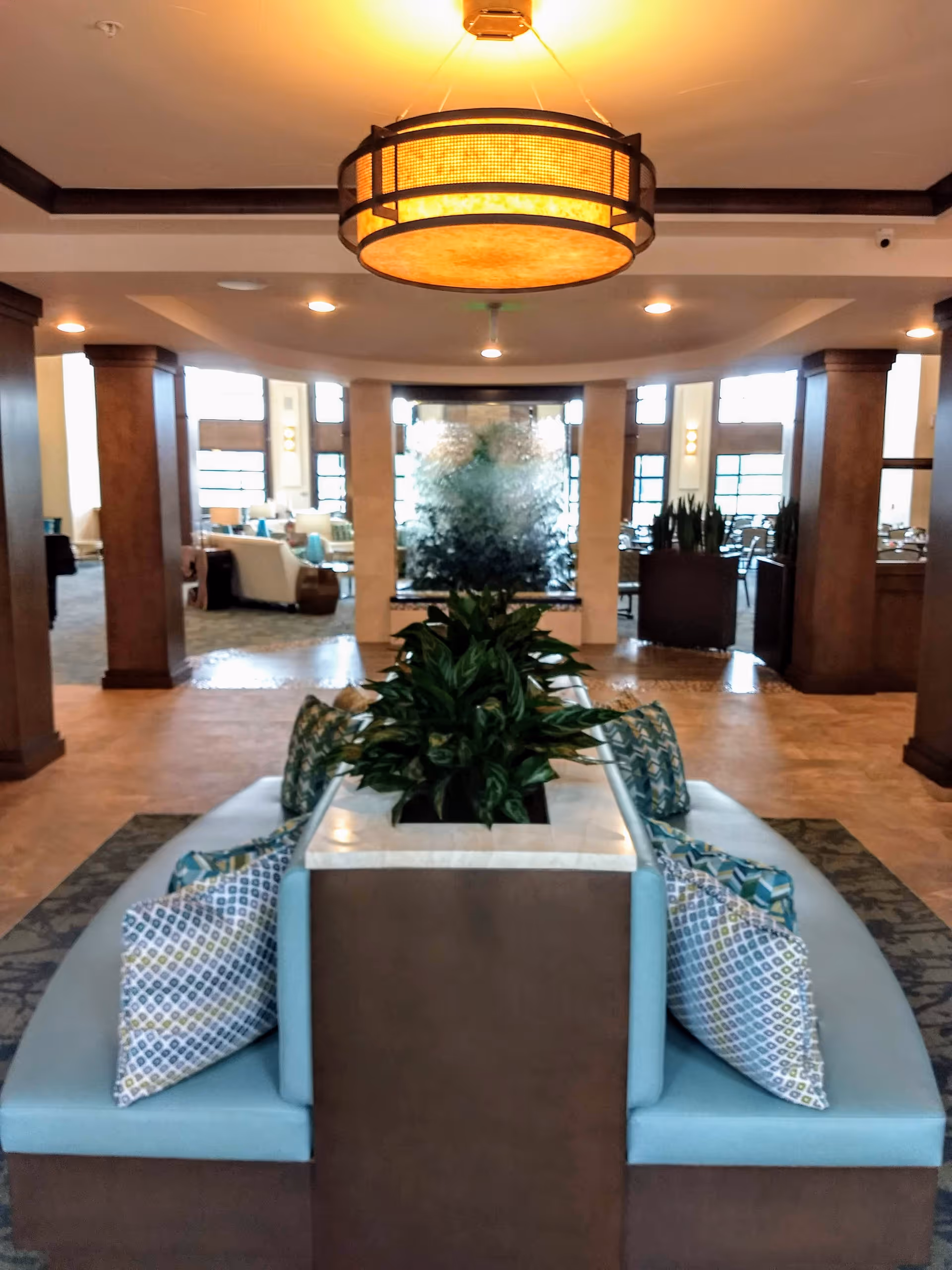 A senior living lobby with circular bench seating surrounding a planted centerpiece, wooden columns, and a large hanging light fixture.