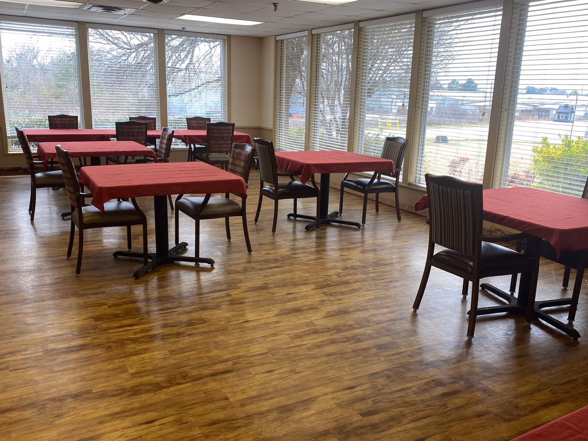 A dining room with several tables covered in red tablecloths and wooden chairs arranged around them. Large windows with blinds allow natural light to fill the room, and the floor is made of wood.
