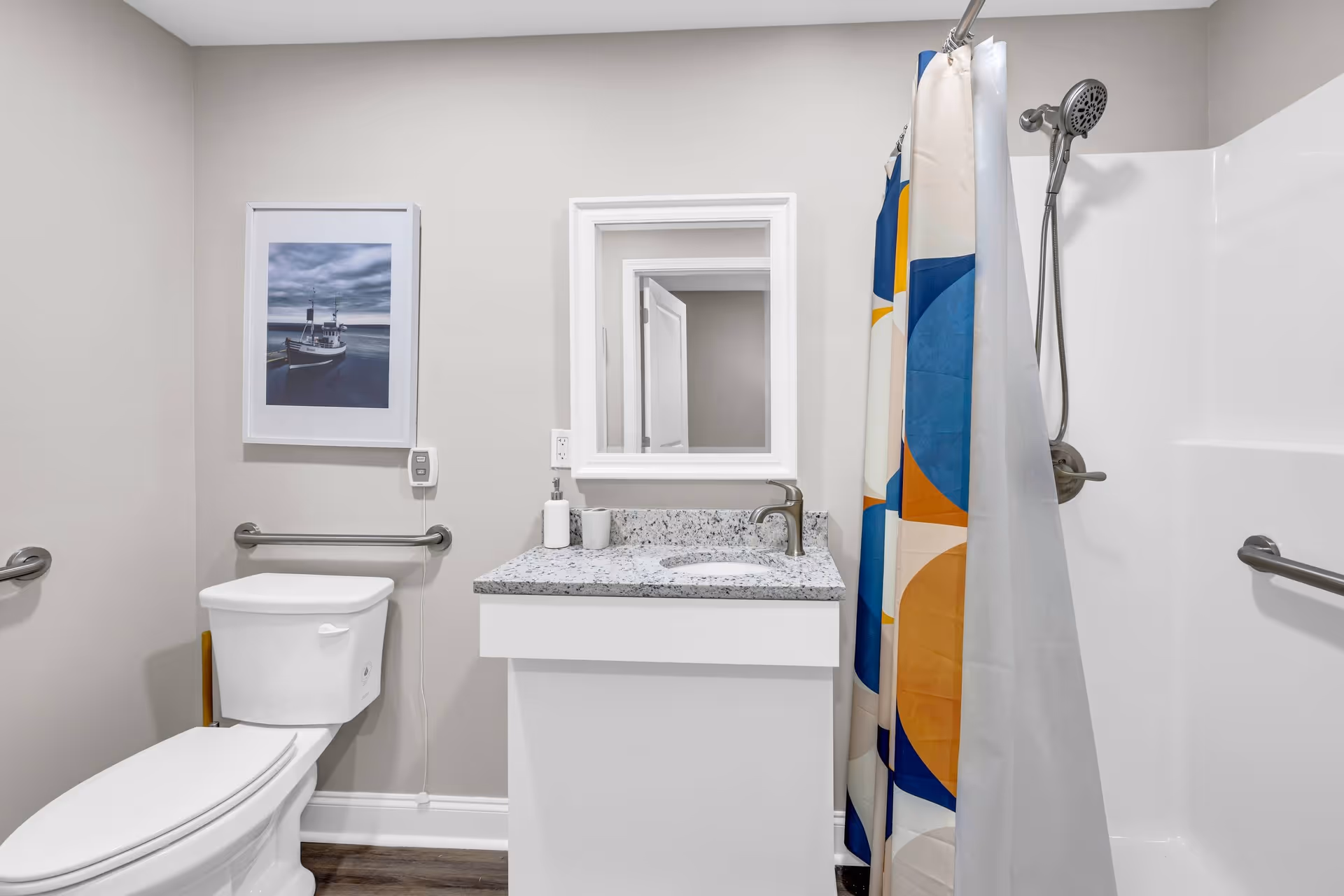 Bright accessible bathroom with a toilet, vanity and mirror, shower with a colorful curtain, and multiple grab bars.