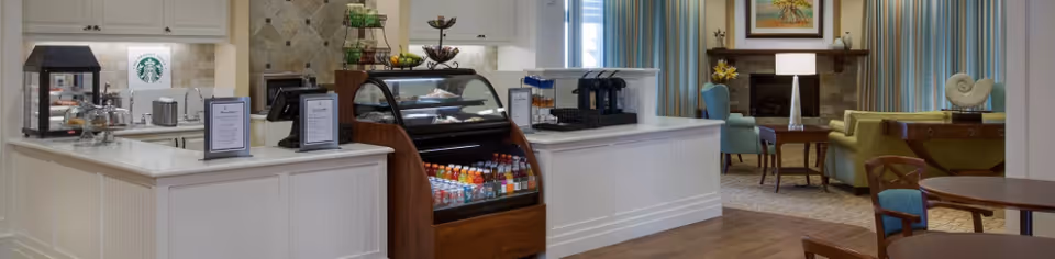 Interior view of a senior living facility's café area with a white counter displaying a variety of beverages in a refrigerated case, coffee machines, and pastries under glass covers. The background shows a cozy seating area with armchairs, a sofa, a lamp, and a fireplace with artwork above it.