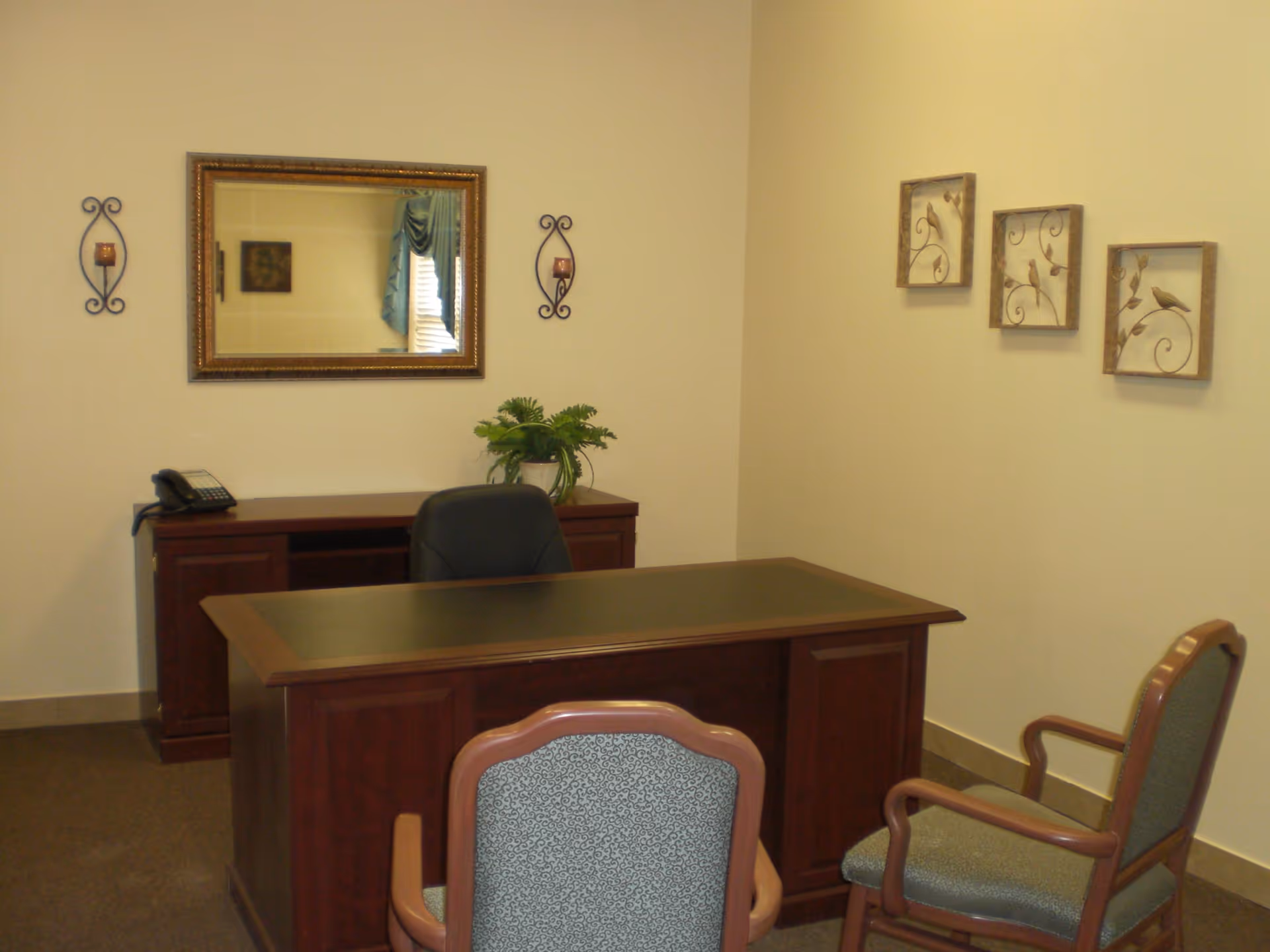 A small furnished office with a wooden desk, two guest chairs, a potted plant, telephone, and framed wall decor.