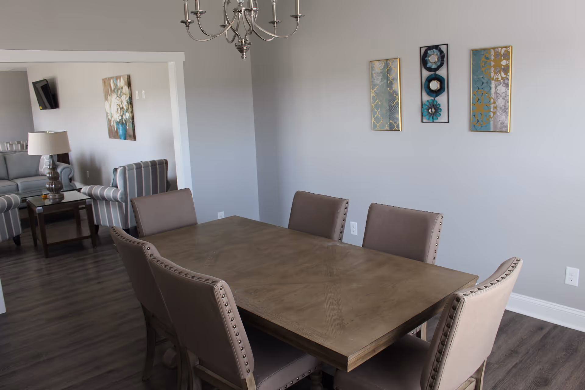 A dining room with a rectangular wooden table surrounded by six upholstered chairs. The walls are light gray with three decorative wall hangings. A chandelier hangs above the table. In the background, there is a living room area with a striped armchair, a gray sofa, a side table with a lamp, and a wall-mounted TV.