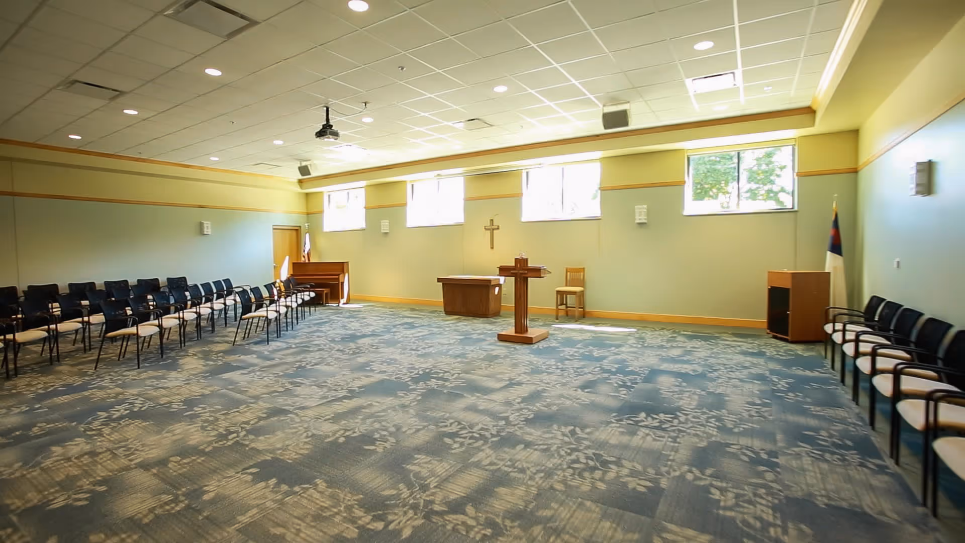 A spacious room with rows of chairs arranged along the left and right walls, a wooden podium in the center, a small table and chair against the far wall, and a piano in the back left corner. The room has large windows near the ceiling letting in natural light, a patterned carpet, and a cross mounted on the wall, suggesting it is a chapel or meeting room.