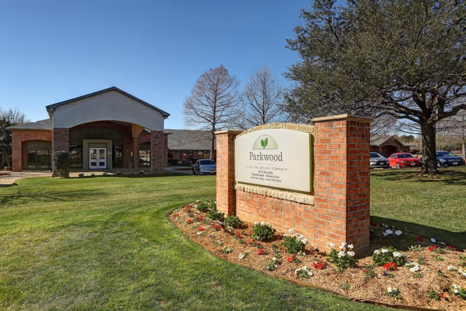 Front entrance of Parkwood Healthcare with a brick sign on a landscaped lawn, the building entryway and parked cars visible behind it.