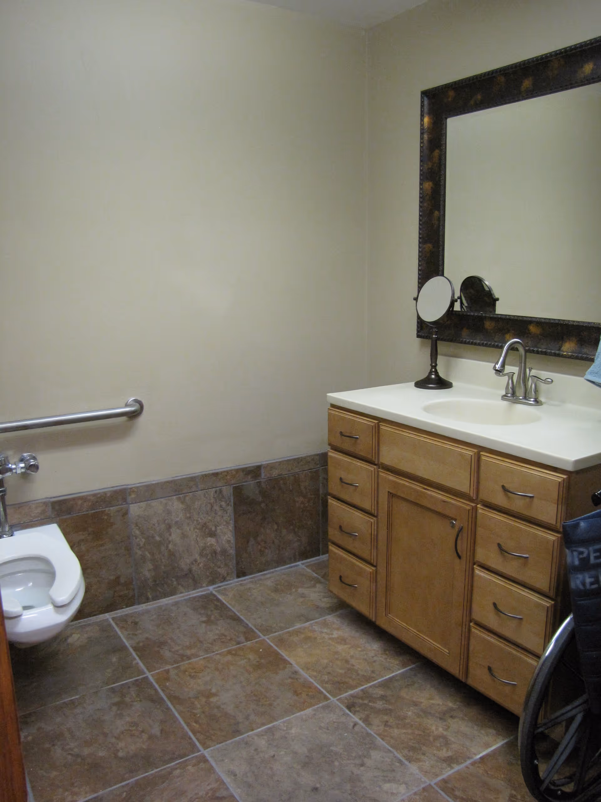 Accessible bathroom with a vanity and mirror, toilet with grab bar, tiled floor, and a wheelchair partially visible.