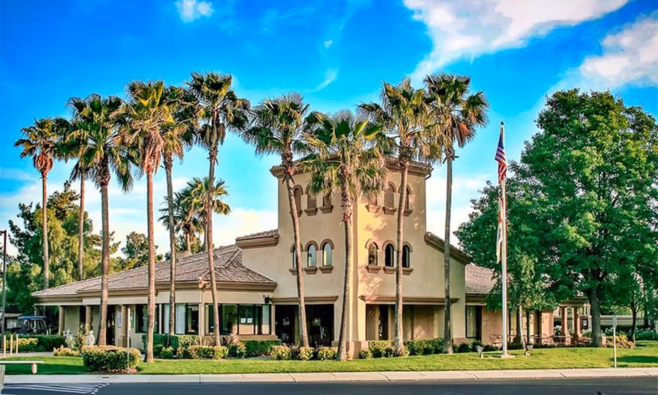 Exterior front view of a Mediterranean-style community building with tall palm trees and an American flag.