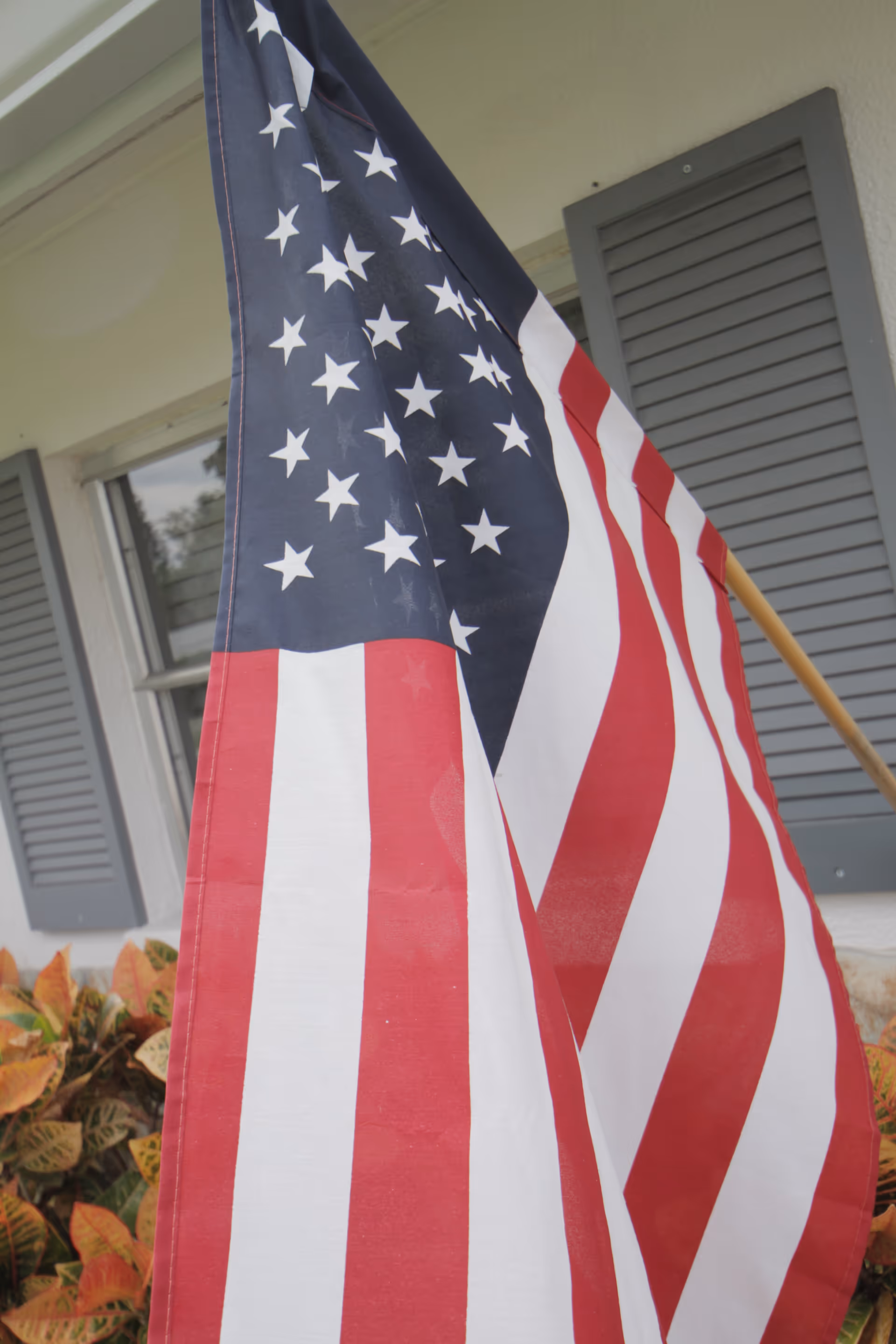 Close-up view of an American flag mounted on a pole in front of a house with gray shutters and a window, with some plants visible near the base of the wall.