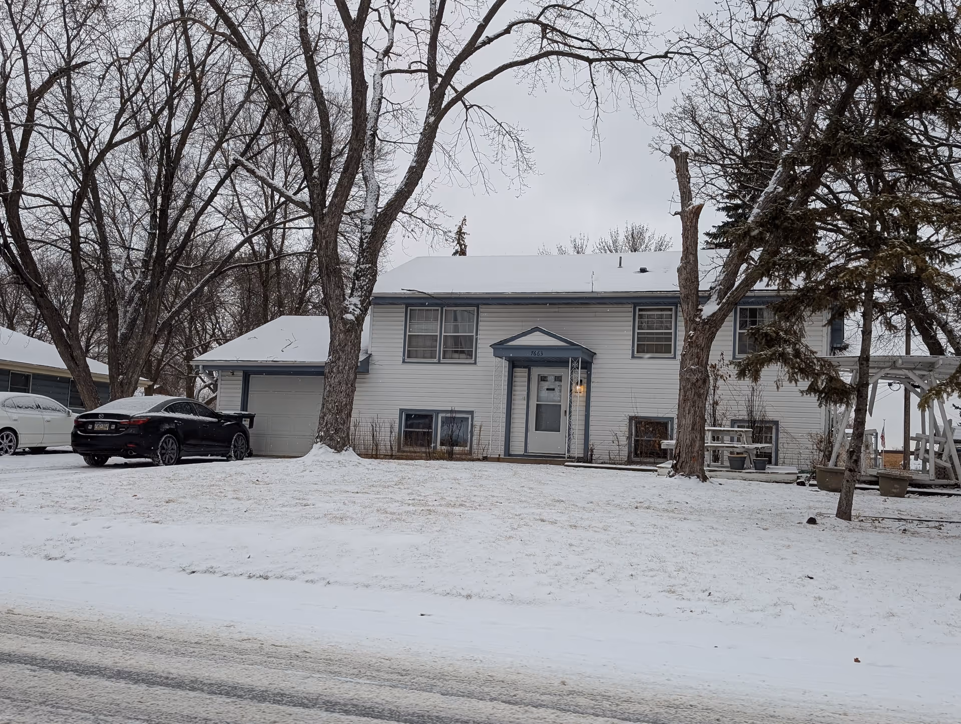 A two-story white house with blue trim and a snow-covered roof and yard. There are several leafless trees in front of the house, a black car parked in the driveway, and a white car partially visible on the left. The sky is overcast and the ground is covered with snow.