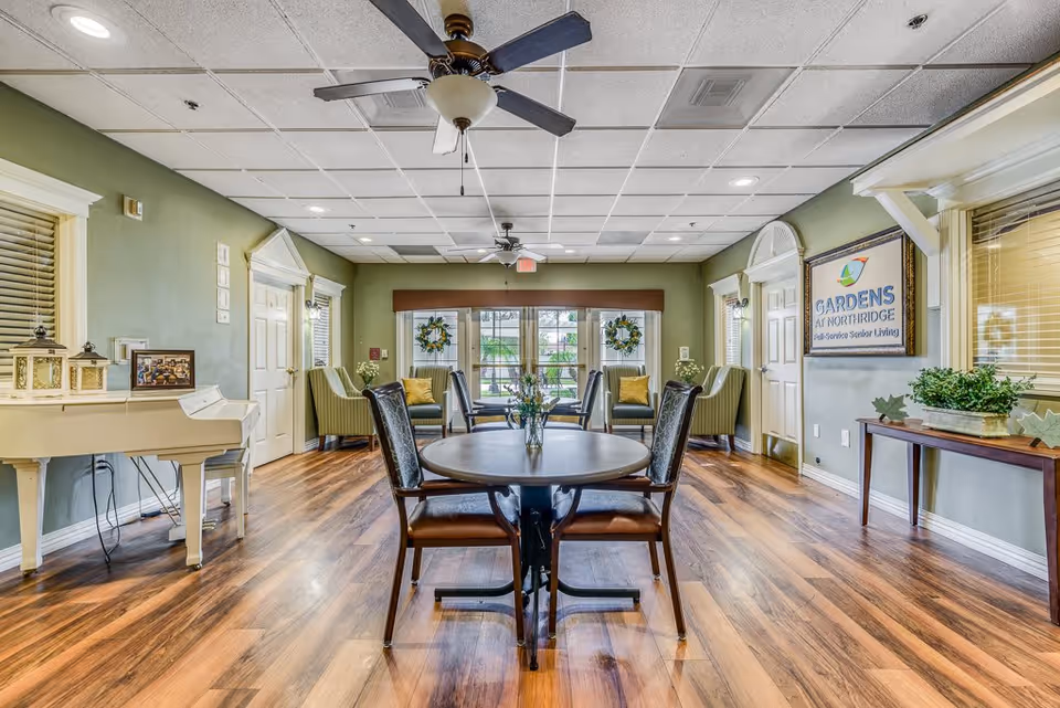 Interior view of a senior living facility common area with wooden flooring, a round table with four chairs in the center, green armchairs along the back wall, ceiling fans, and large windows with wreath decorations. A white piano is on the left side, and a sign on the right wall reads 'Gardens At Northridge'.
