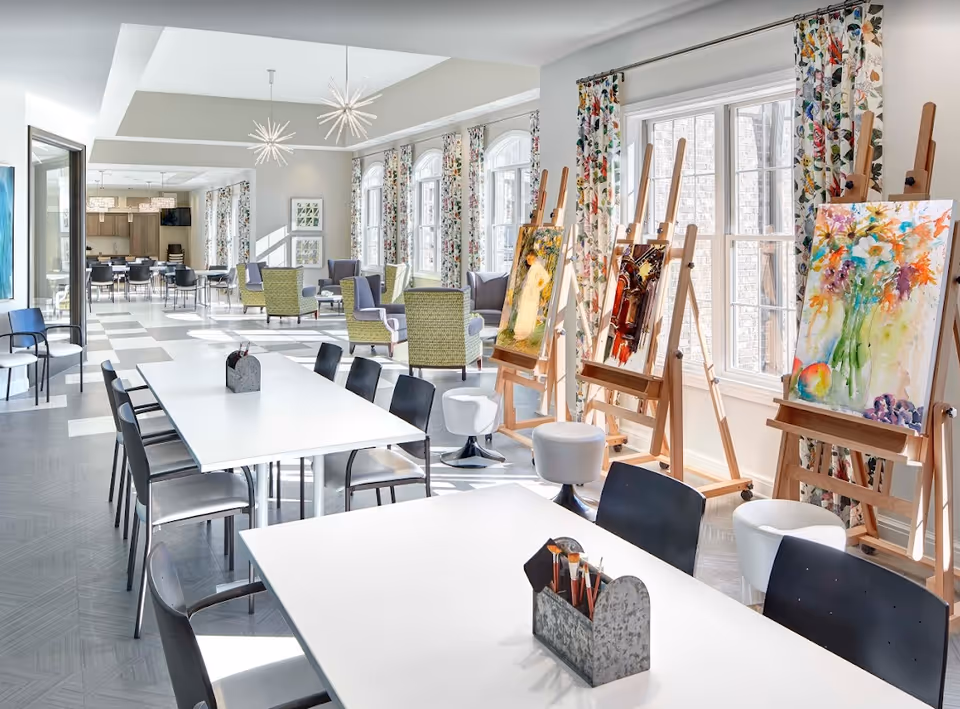Bright communal art studio with long tables and chairs, easels displaying colorful paintings, and lounge seating by large windows.