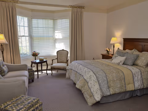 A cozy bedroom with a large bed covered in patterned bedding, a wooden headboard, two bedside tables with lamps and flowers, a sitting area with a sofa, an armchair, a small table, and a large bay window with blinds and curtains.