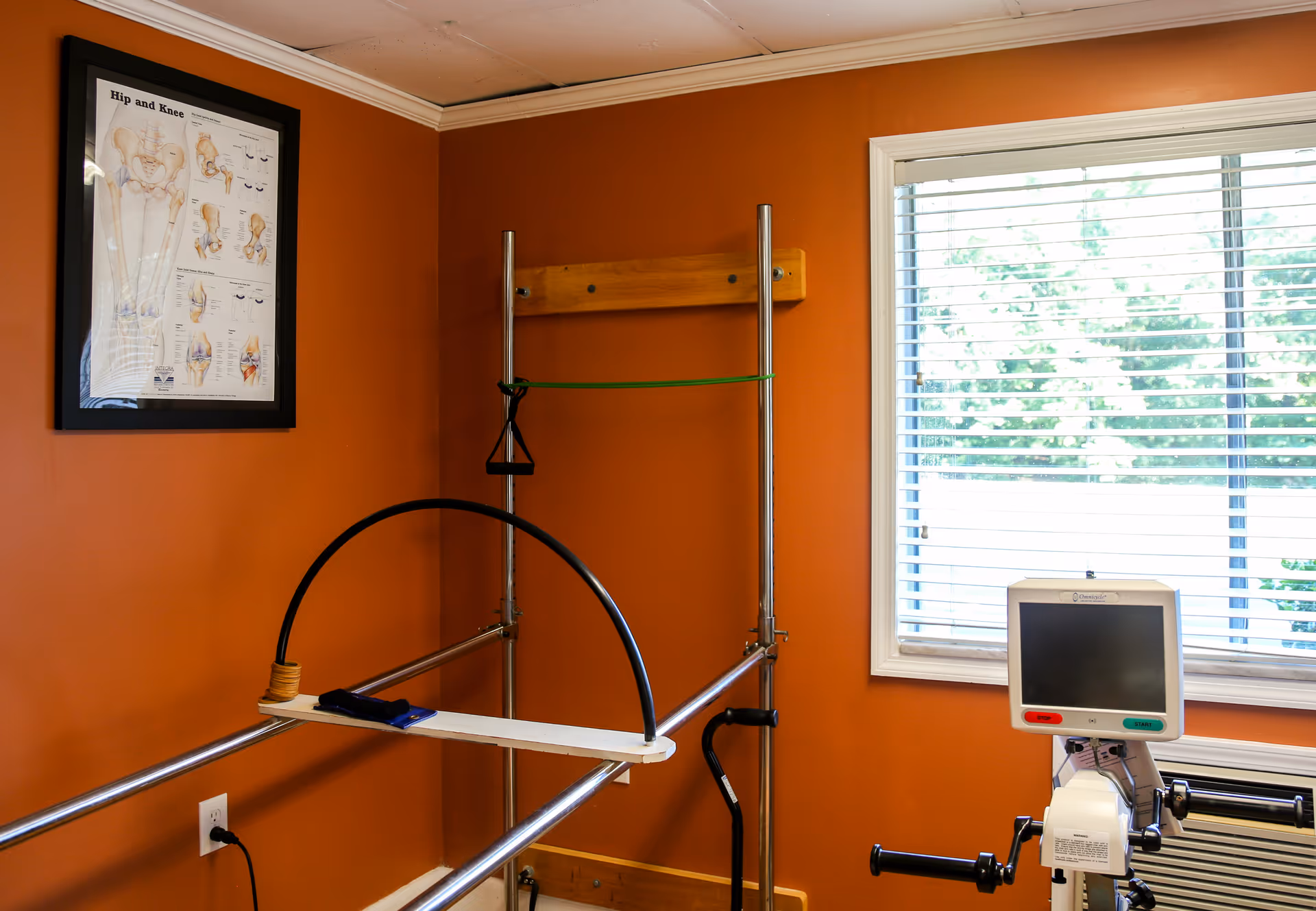 A small physical therapy room with orange walls featuring exercise equipment including a hand cycle machine and a metal frame with resistance bands. A framed anatomical chart of hip and knee joints hangs on the wall, and a window with white blinds lets in natural light.