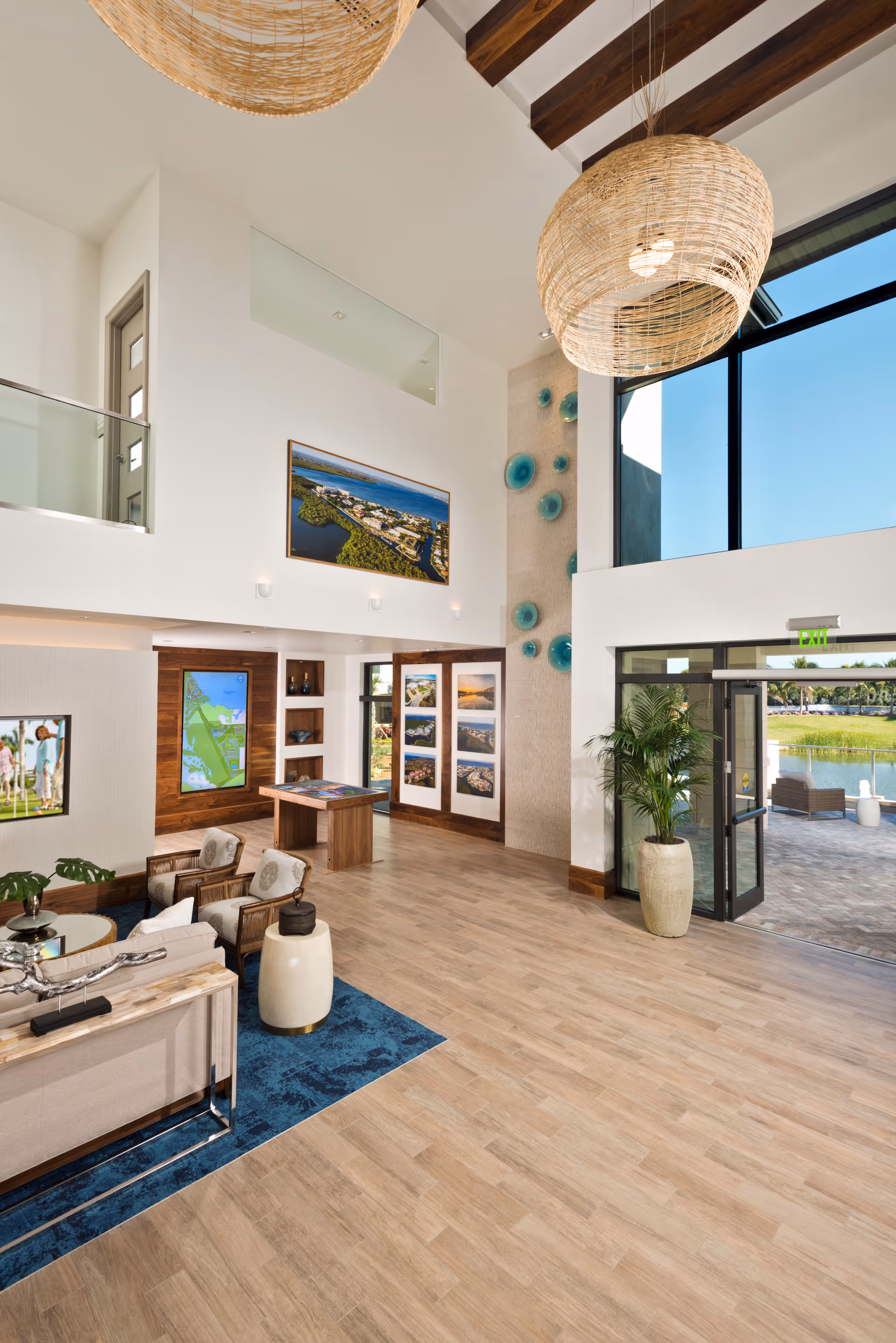 Bright and spacious interior of the Shell Point Welcome Center featuring high ceilings with wooden beams, large windows letting in natural light, modern wicker pendant lights, a seating area with chairs and a sofa on a blue rug, a wooden table with maps and pictures on the walls, and a glass door leading outside to a patio with water views.