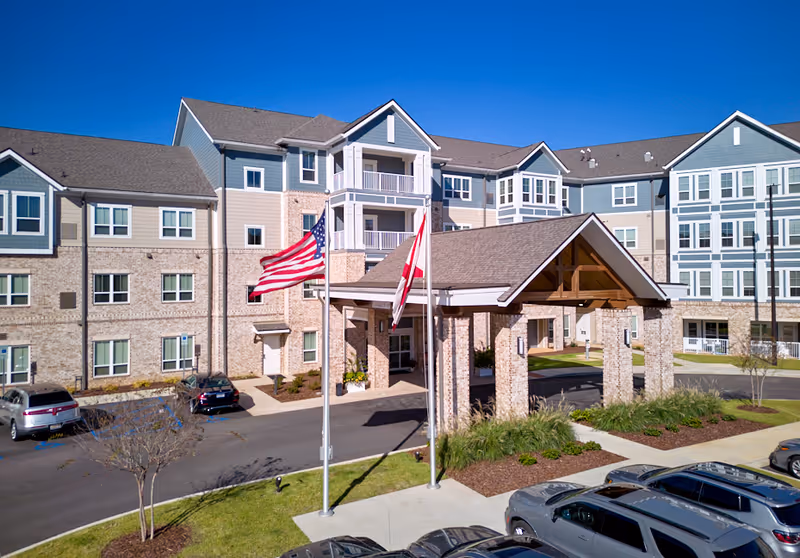 Exterior view of a multi-story senior living facility with a covered entrance supported by stone pillars. Two flagpoles with the American flag and another flag are positioned in front of the entrance. Several cars are parked in the parking lot, and the building features a combination of brick and siding with multiple windows under a clear blue sky.