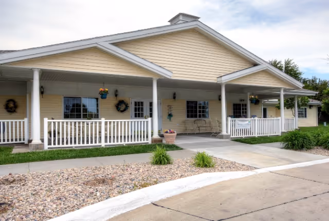 Exterior view of a single-story senior living facility building with beige siding, white trim, and a covered porch supported by white columns. The porch has hanging flower baskets, decorative wreaths on the walls, and a bench. There is a paved walkway and landscaped area with rocks and small plants in front of the building.