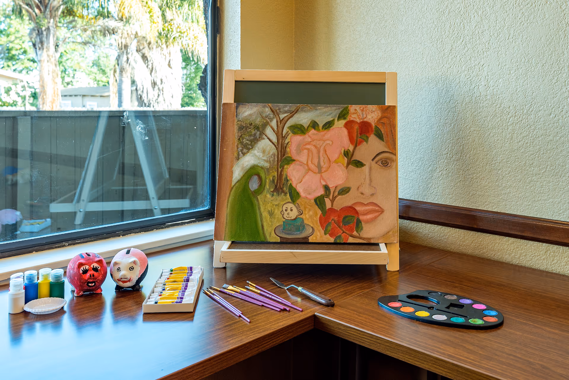 An art easel displaying a floral face painting with paint palettes, brushes, and small painted figures arranged on a wooden table by a window.