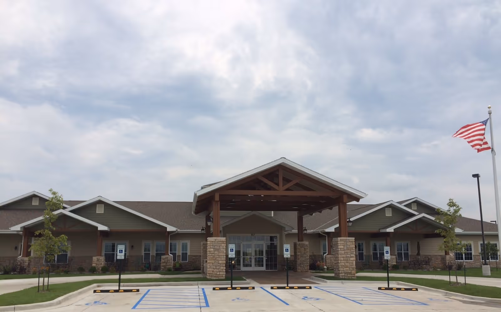 Front exterior view of Oak Pointe of Maryville facility with a covered entrance supported by stone pillars, multiple handicap parking spaces in front, small trees planted along the walkway, and an American flag on a flagpole to the right under a cloudy sky.