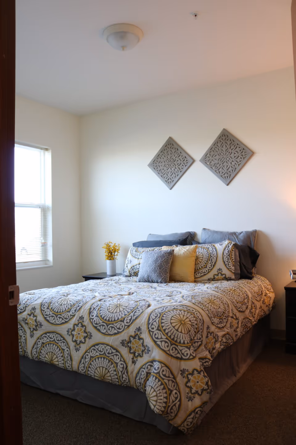 A neatly made bed with patterned bedding in shades of gray, yellow, and white in a bedroom. There are multiple pillows arranged on the bed. On the wall above the bed are two decorative square wall hangings with intricate designs. A window with blinds is on the left side, and a small bedside table with a vase of yellow flowers is next to the bed. The room has light-colored walls and a ceiling light fixture.