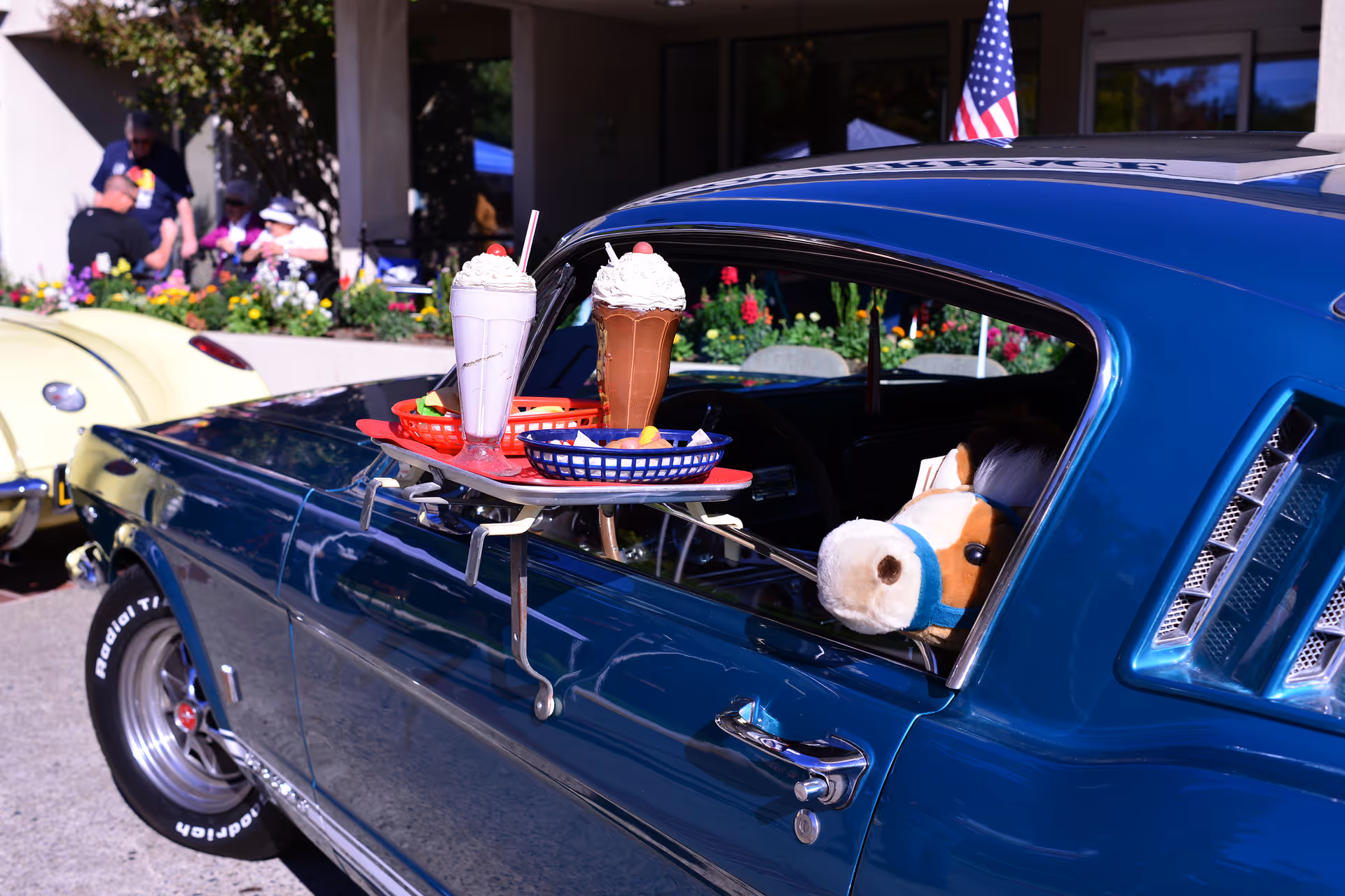 A blue vintage car with a tray attached to the open window holding two milkshakes and a basket of fries. A stuffed horse toy is visible inside the car. In the background, people are sitting near a flower bed outside a building with an American flag on the car roof.