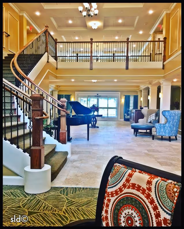 Spacious and elegant senior living facility interior featuring a grand staircase with wooden handrails and black iron balusters, a black grand piano in the center, comfortable seating including a blue patterned armchair and a cushioned chair with a colorful circular pattern, beige tiled flooring, and large windows with blue curtains allowing natural light to fill the room.