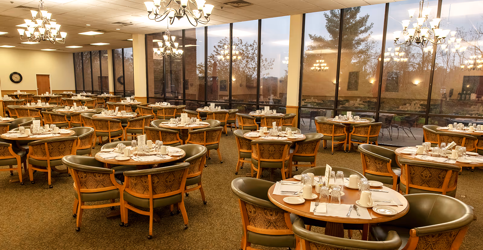 A spacious dining room with multiple round wooden tables set with white cups, plates, utensils, and napkins. Each table is surrounded by cushioned chairs with wooden frames and patterned backs. Large floor-to-ceiling windows line one side of the room, showing an outdoor patio and trees outside. Elegant chandeliers hang from the ceiling, illuminating the room warmly.