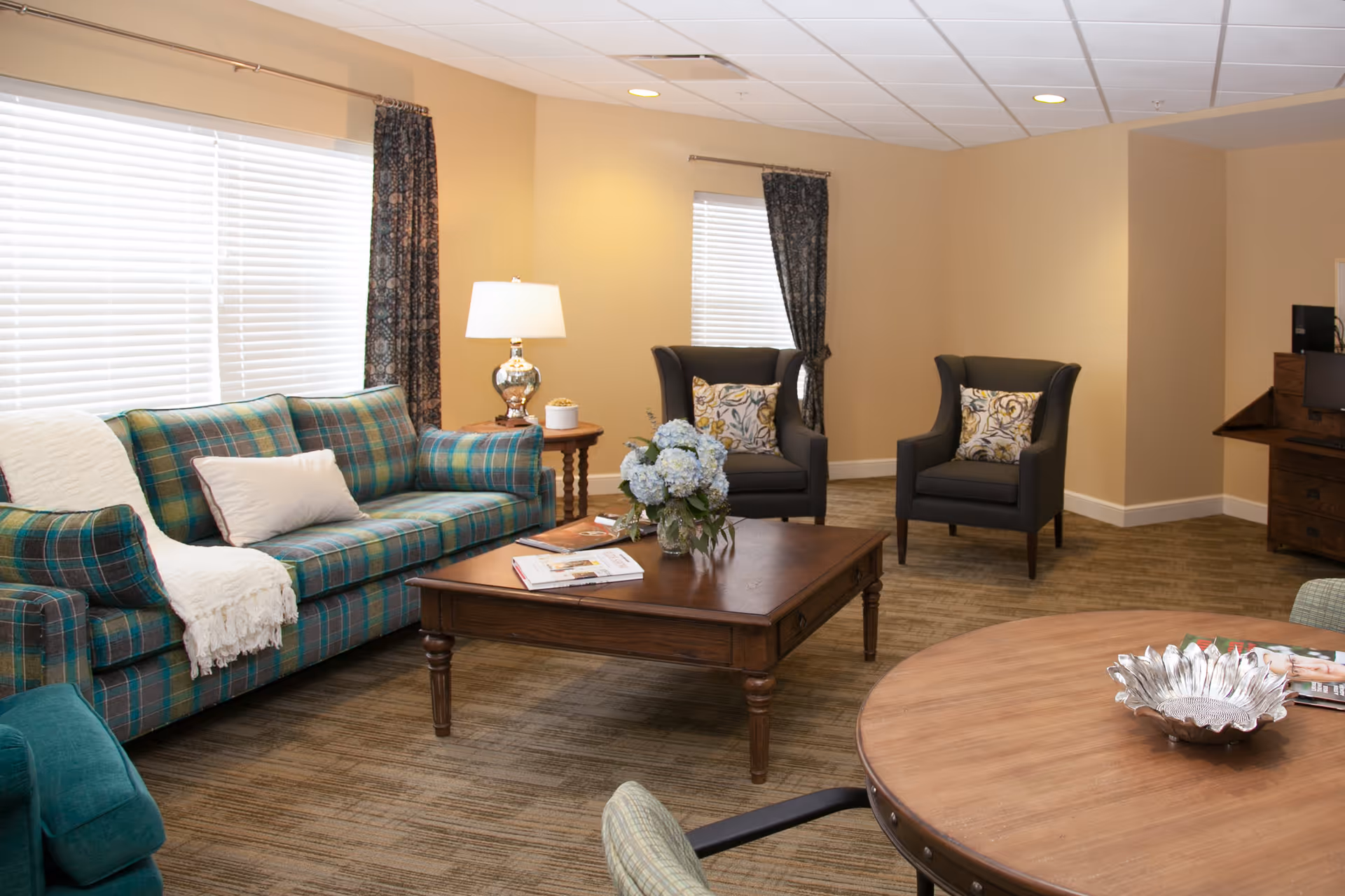 A cozy living room with a plaid sofa, two dark armchairs with patterned cushions, a wooden coffee table with a flower arrangement and magazines, a round wooden table with a decorative bowl, and a side table with a lamp. The room has beige walls, carpeted floor, and windows with blinds and dark curtains.