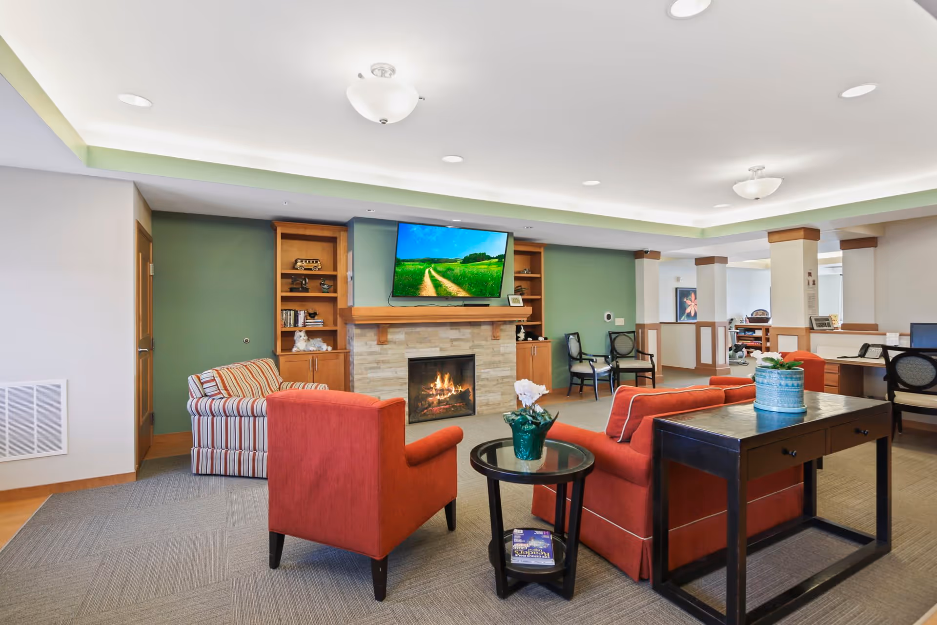 A cozy senior living facility common area with a fireplace and a wall-mounted TV above it. The room features comfortable seating including a red armchair, a red sofa, and a striped armchair. There are built-in wooden shelves flanking the fireplace, a small round glass table with a green vase and white flowers, and a dark wooden console table with a blue pot and plant. The ceiling has recessed lighting and a modern light fixture, and the walls are painted green and white.