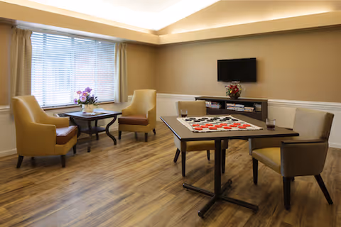 A bright common room with armchairs, a game table arranged for checkers, a wall-mounted TV, and a small table by the window.