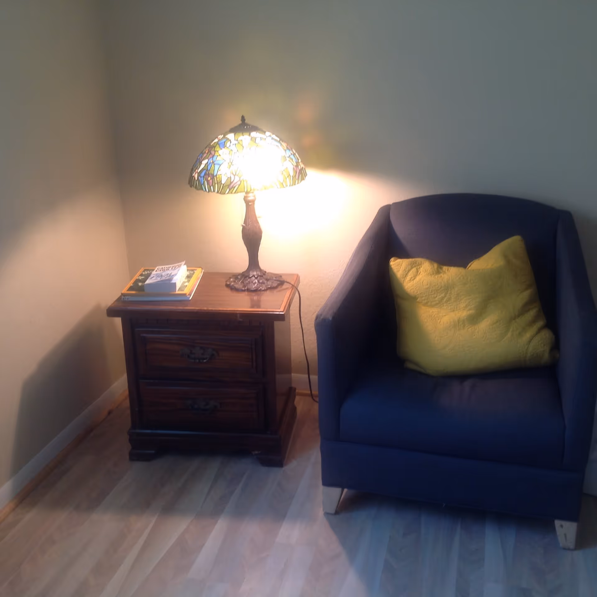 A cozy corner with a lit stained-glass table lamp on a wooden side table beside a blue armchair with a yellow pillow.
