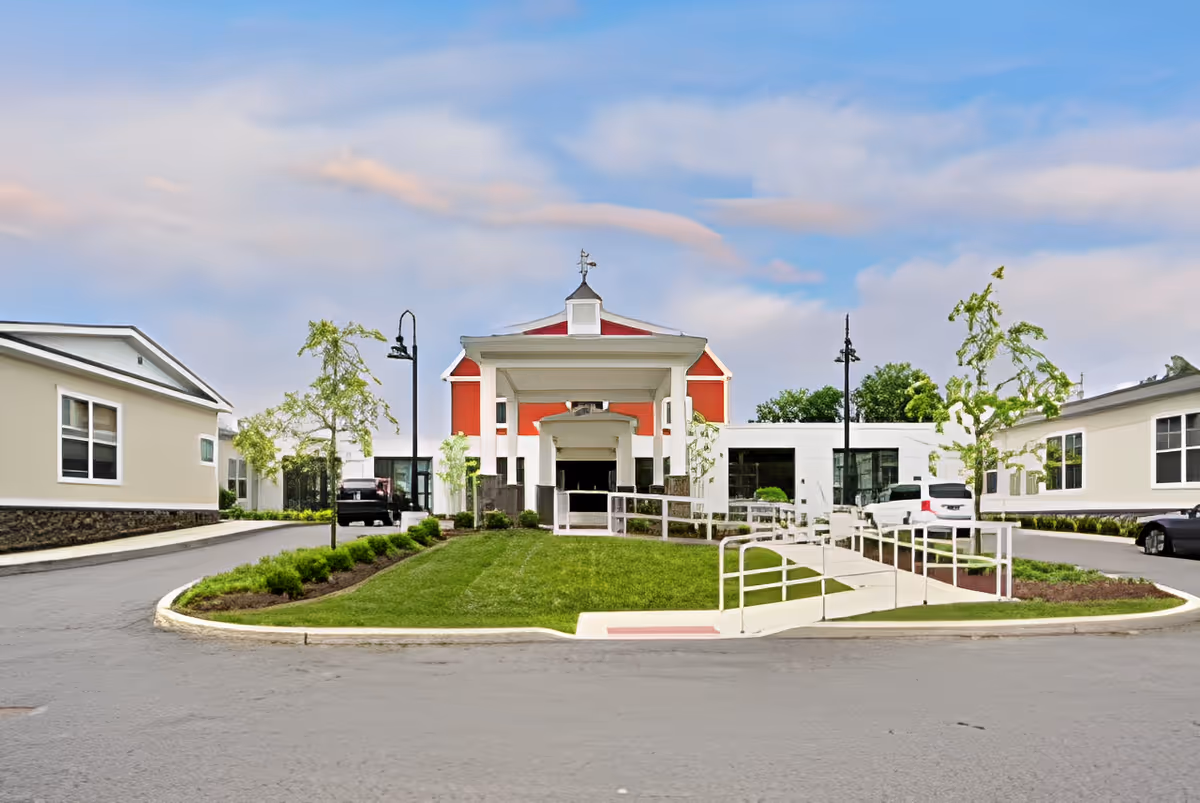 Front exterior view of Church Hill Village facility with a central entrance featuring a covered portico, flanked by two single-story buildings, landscaped greenery, small trees, and a paved driveway with parked cars under a partly cloudy sky.