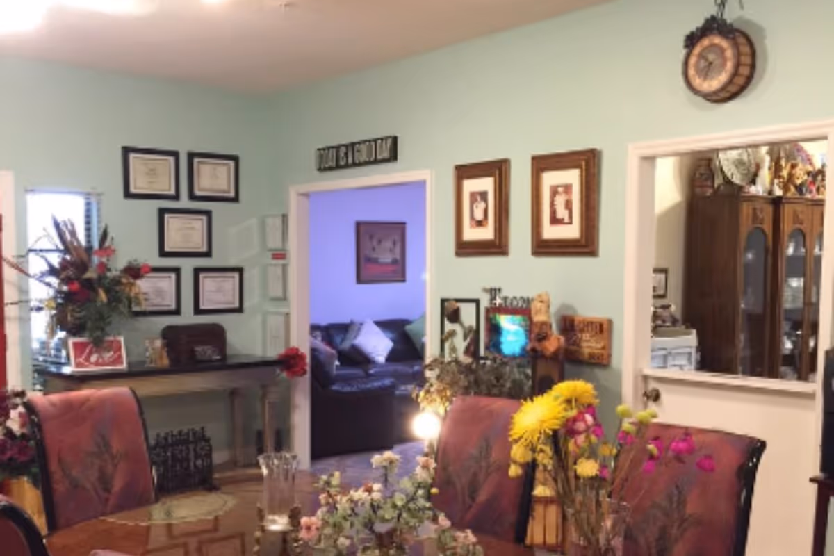 Dining area with a table and floral centerpieces, upholstered chairs, and framed pictures on mint-green walls looking into a living room.
