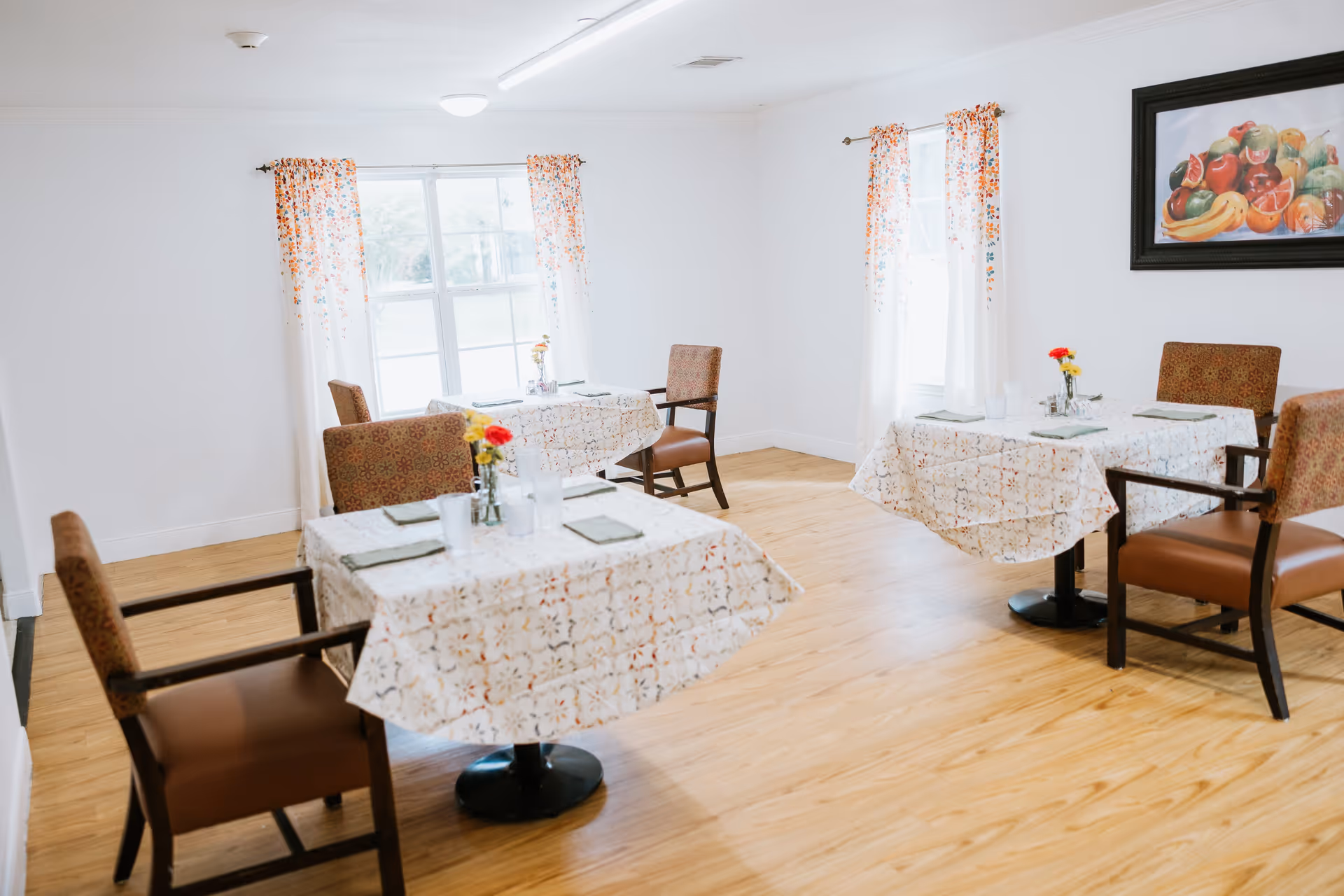 A bright dining room with three tables covered in patterned tablecloths, each set with napkins, cups, and small flower vases. The room has wooden flooring, white walls, two windows with floral curtains, and a framed painting of assorted fruits on the wall.