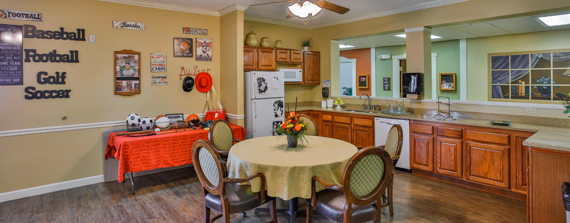 Communal dining area and kitchenette with a round table and chairs, wooden cabinets, and a sports-themed display on the wall.