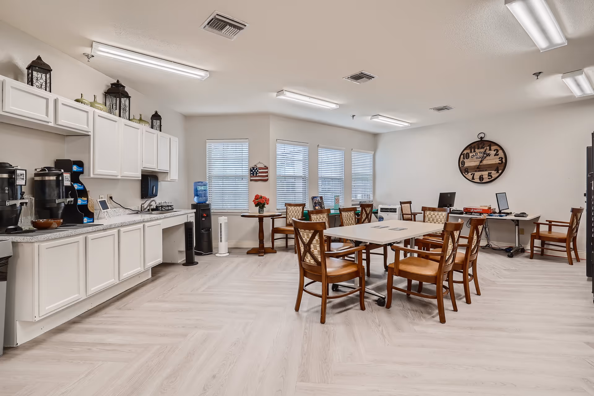 A bright, spacious room with white walls and light wood flooring featuring a kitchenette area with white cabinets, a coffee station, and a water cooler on the left. In the center, there are tables and wooden chairs with cushioned seats arranged for dining or activities. On the right side, there are desks with computer monitors and chairs, and a large wall clock is mounted on the wall.