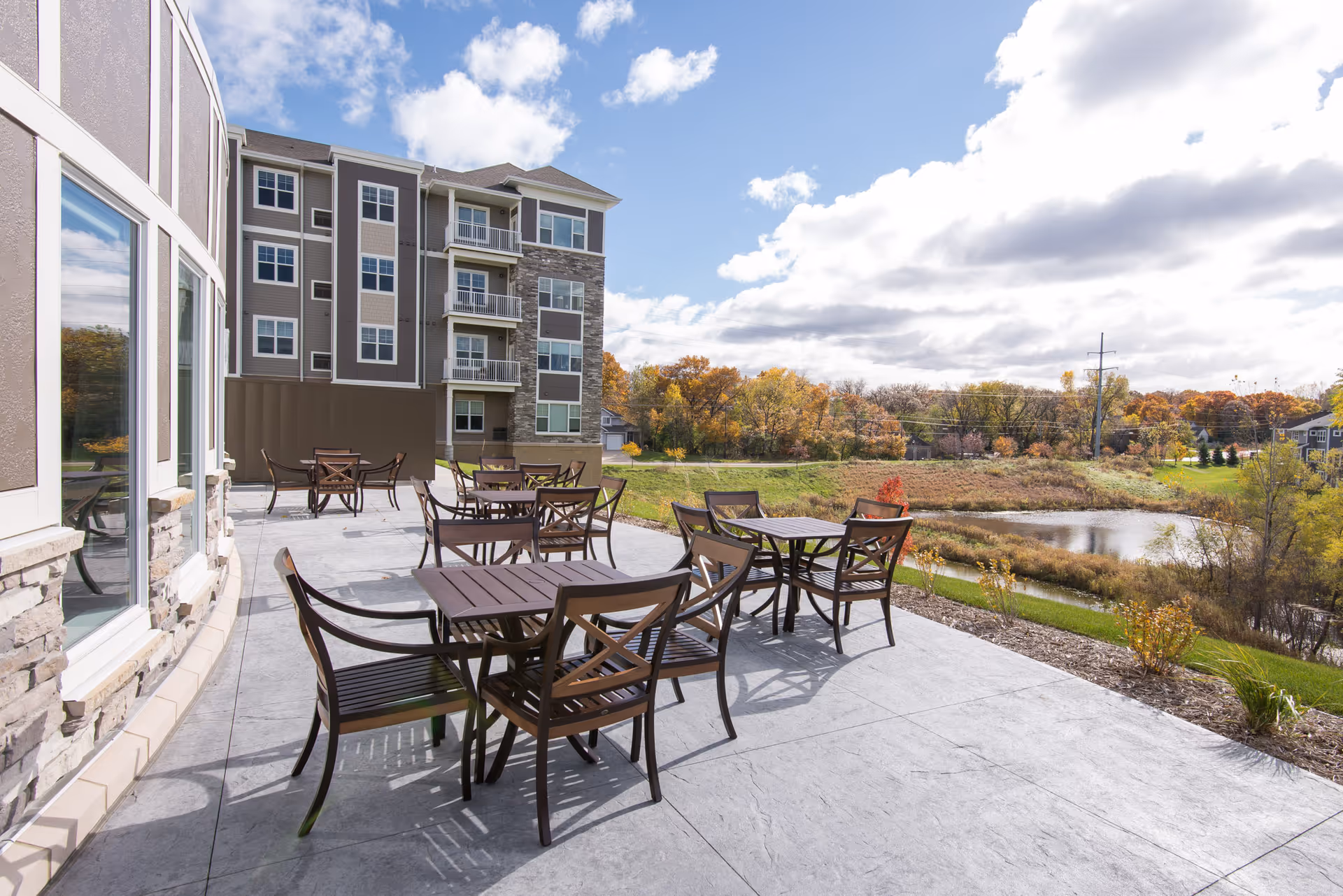 Outdoor patio area at Stonehaven Senior Living with several wooden tables and chairs arranged on a concrete surface. The patio overlooks a scenic view of a pond, grassy area, and trees with autumn foliage under a partly cloudy sky. The building exterior features large windows and stone accents.