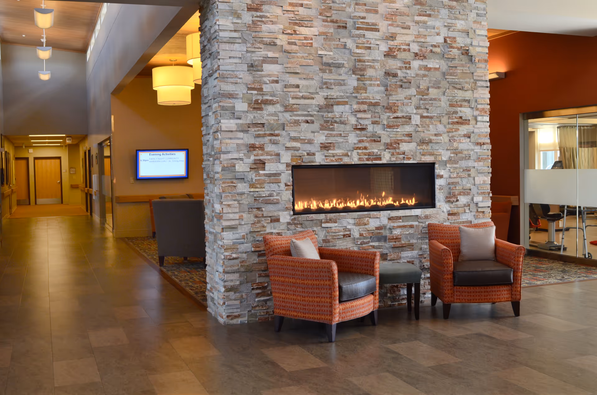 A cozy seating area in a senior living facility with two orange patterned armchairs and a small ottoman in front of a modern stone fireplace with a long horizontal flame. The area is well-lit with ceiling lights and has a hallway leading to other rooms. There is a digital screen on the wall displaying evening activities and a glass-walled room visible on the right side.