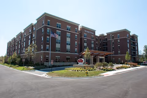 Exterior view of a large multi-story brick building with the sign 'Victory Centre' visible near the entrance. The building is surrounded by landscaped greenery, a driveway, and an American flag flying on a flagpole.