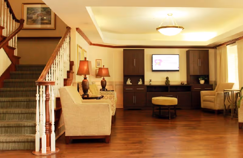 A cozy living room area in an assisted living facility featuring wooden flooring, beige armchairs, a small round ottoman, two table lamps on side tables, a staircase with carpeted steps and white railings, and a wall-mounted television flanked by dark wooden cabinets.