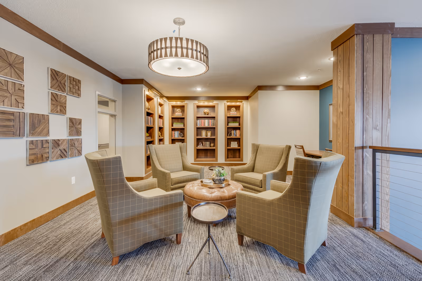 A cozy sitting area with four beige armchairs arranged around a round leather ottoman in the center. Behind the chairs, there are built-in wooden bookshelves filled with books and decorative items. The room features a modern ceiling light fixture, wooden trim, and a carpeted floor. On the left wall, there is a decorative wooden wall art installation. To the right, a wooden column and railing with metal cables are visible.