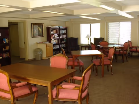 A bright common room with several wooden tables and upholstered chairs, bookshelves, armchairs, and windows letting in daylight.
