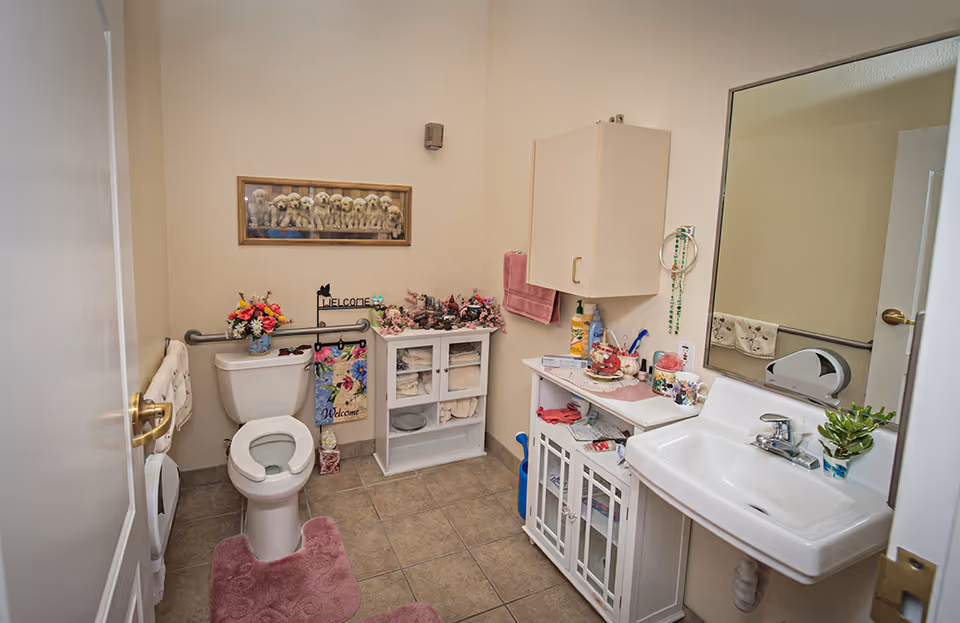 Small, decorated bathroom featuring a toilet, sink with mirror, storage cabinets, and floral decorations.
