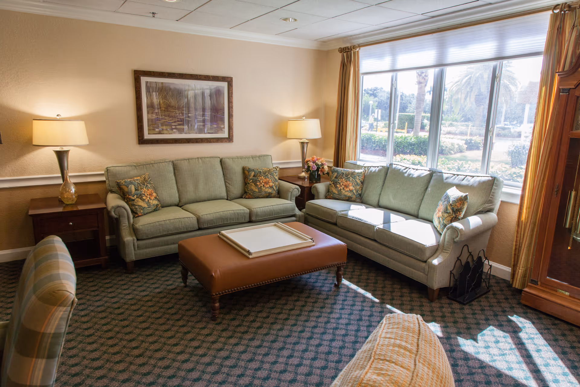 A cozy living room with two light green sofas adorned with floral cushions, a brown leather ottoman in the center, two side tables each with a lamp, a large window with yellow curtains letting in natural light, a framed picture on the wall, and a patterned carpeted floor.