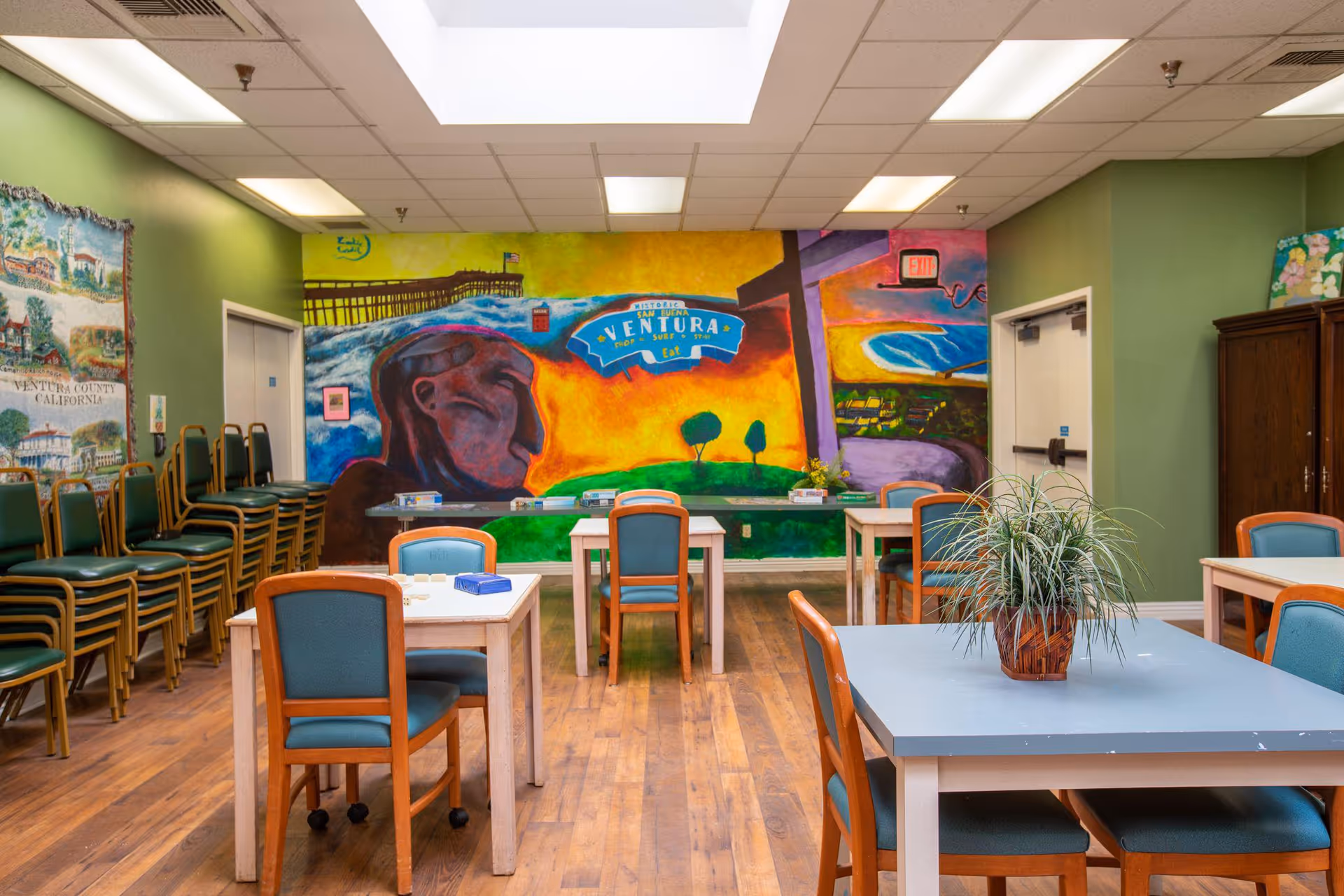 A brightly lit room with several tables and chairs arranged for group seating. The walls are painted green and feature colorful murals, including one with the word 'Ventura' and artistic depictions of landscapes and faces. There are stacks of additional chairs against one wall and a potted plant on one of the tables. The room has a wooden floor and a drop ceiling with fluorescent lights.