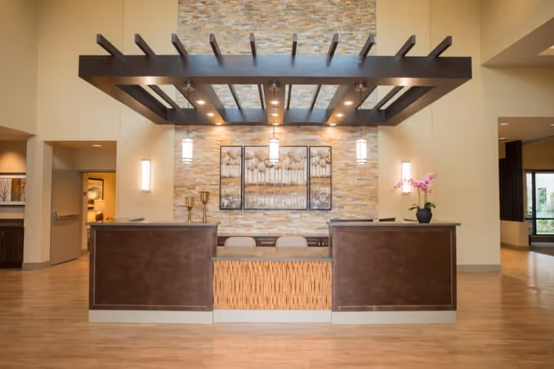 A modern reception desk area in a senior living facility with a wooden floor, a stone accent wall behind the desk, three pendant lights hanging from a dark wooden beam structure, and a triptych artwork featuring abstract trees. There are two chairs behind the desk and a small potted orchid on the right side.