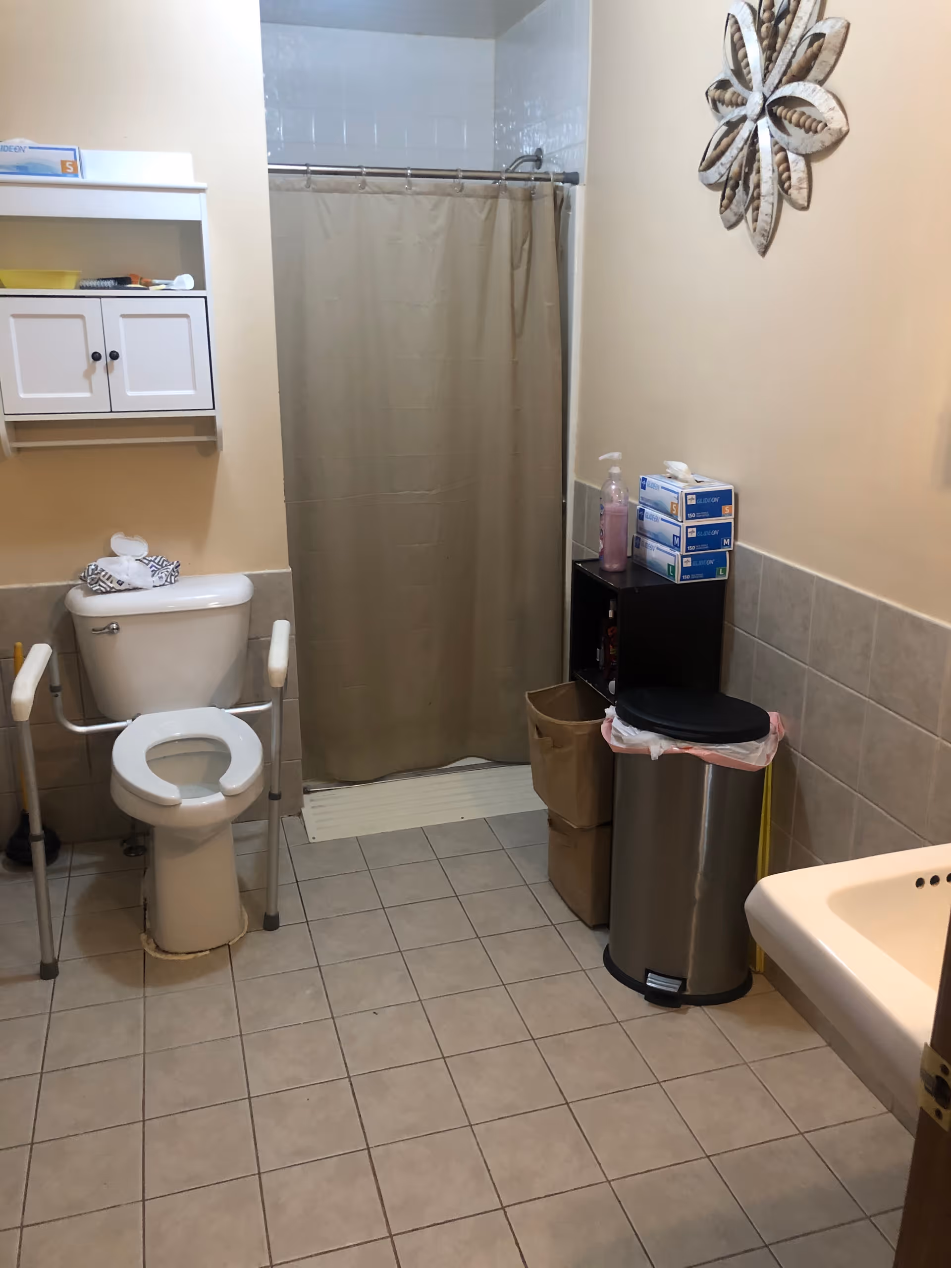 A bathroom with a toilet equipped with safety rails, a shower with a beige curtain, a wall-mounted cabinet, a trash can with a plastic liner, a small shelving unit with boxes of gloves and a bottle of soap, and a sink partially visible on the right. The walls are tiled halfway up and painted beige above.
