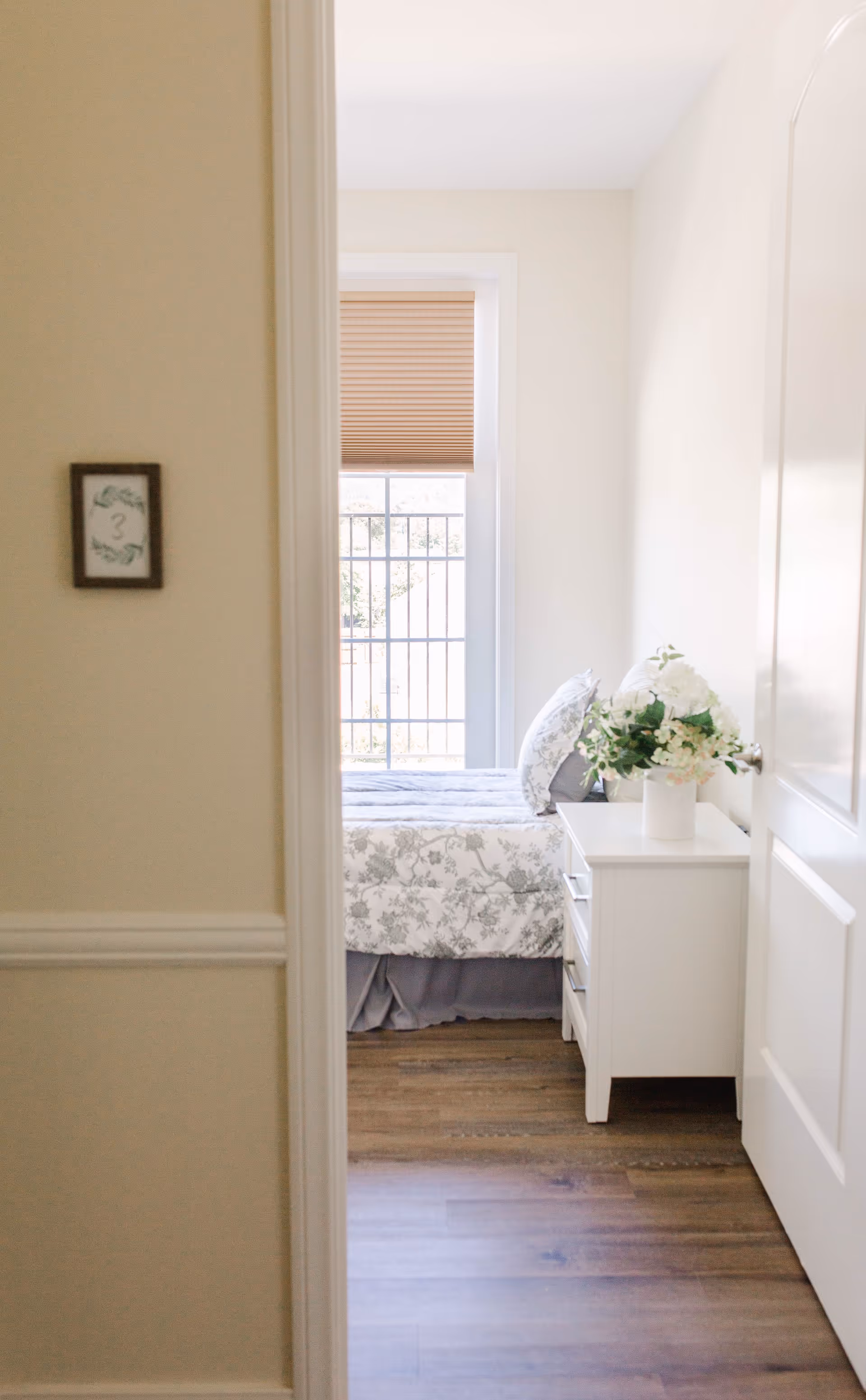 View through a doorway into a bright bedroom with a bed covered in floral bedding, a white nightstand with a vase of white flowers, and a window with a beige blind partially down.
