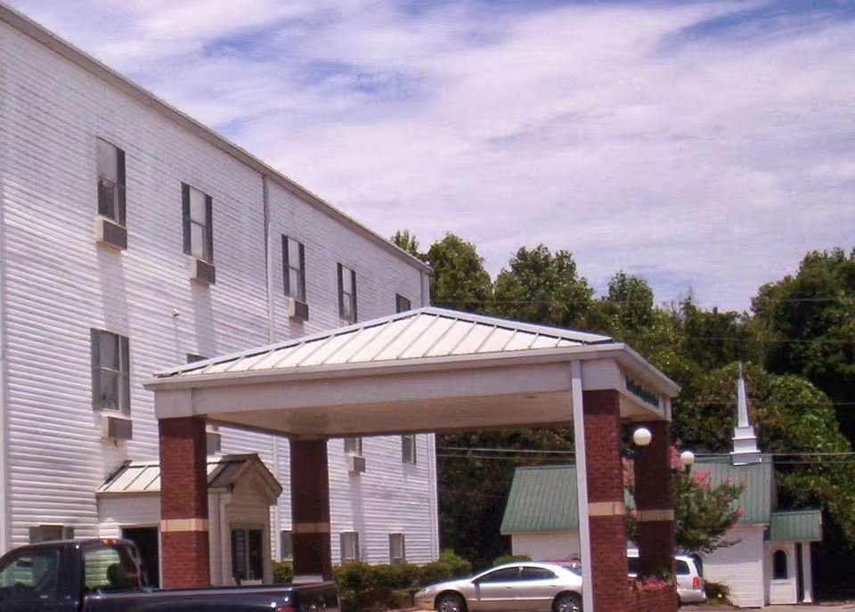 Exterior view of a multi-story white building with multiple windows and air conditioning units. The building has a covered entrance supported by brick columns with a metal roof. Several cars are parked nearby, and there are trees and a small white building with a green roof and a steeple in the background under a partly cloudy sky.